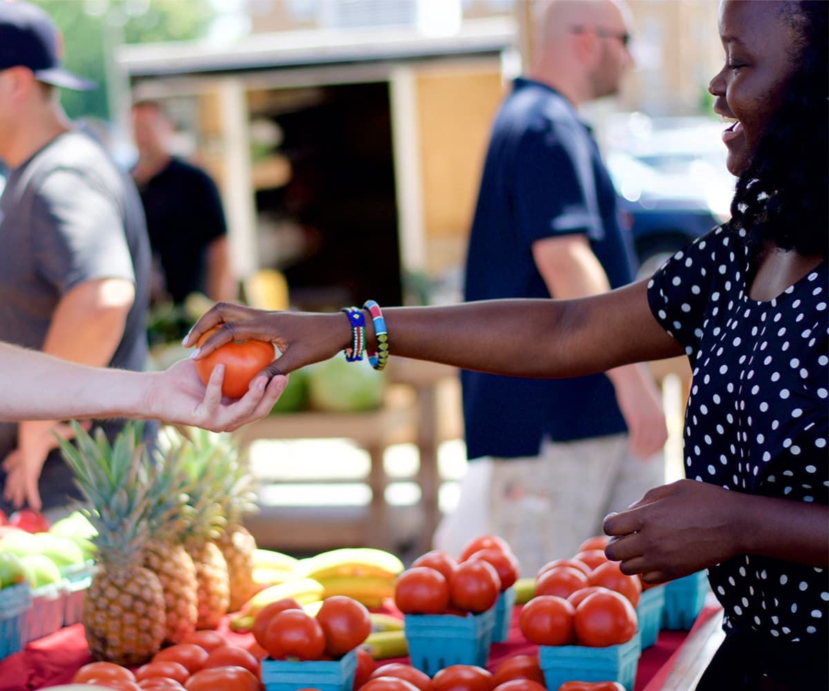 A woman handing a ripe tomato to a customer at a farmers market.