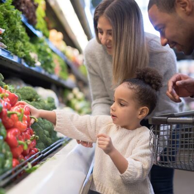 A young family shopping at a grocery store with a young girl selecting a red pepper from the produce shelf.
