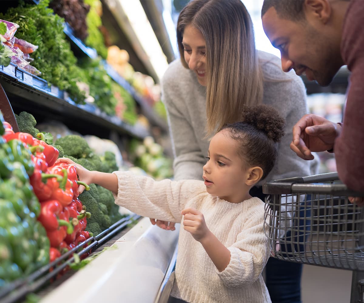 A young family shopping at a grocery store with a young girl selecting a red pepper from the produce shelf.