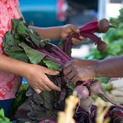 The hands of two women passing a bunch of beets between each-other at a farmers market.