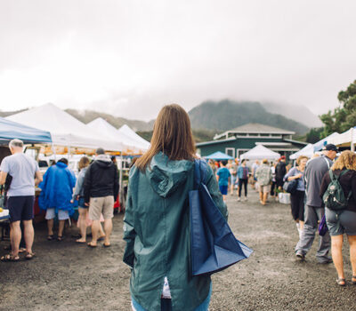 Image of person standing in front of a farmers market wearing a rain coat and carrying a tote bag.