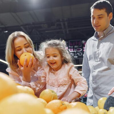 A woman smelling a fresh orange next to a young girl and man standing in a grocery's produce section.